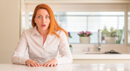Redhead woman at kitchen afraid and shocked with surprise expression, fear and excited face.