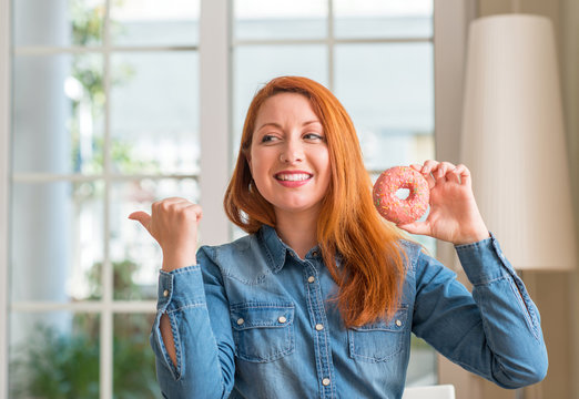Redhead woman holding donut at home pointing with hand and finger up with happy face smiling