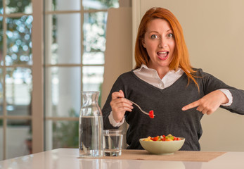 Redhead woman eating fruit bowl, kiwi and strawberry at home very happy pointing with hand and...