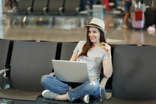 Young smiling traveler tourist woman in hat sit with crossed legs, working on laptop holding bitcoin wait in lobby hall at airport. Passenger traveling abroad on weekends getaway. Air flight concept. - Powered by Adobe