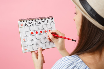 Back rear view of woman in blue dress, hat holding red pencil, female periods calendar for checking menstruation days isolated on pink background. Medical healthcare, gynecological concept. Copy space