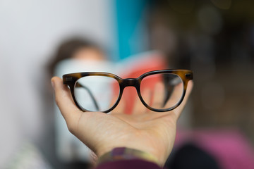 Holding up glasses behind a girl reading a book that is blurry in the background