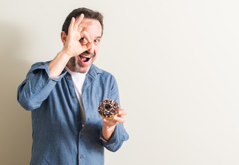 Senior man eating chocolate donut with happy face smiling doing ok sign with hand on eye looking through fingers