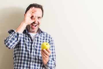 Senior man holding a green apple with happy face smiling doing ok sign with hand on eye looking through fingers