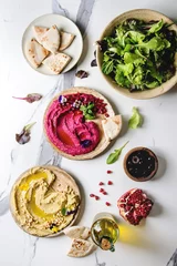 Foto auf Acrylglas Variety of homemade traditional and beetroot spread hummus with pine nuts, olive oil, pomegranate served on ceramic plates with pita bread and green salad on white marble background. Flat lay, space. © Natasha Breen
