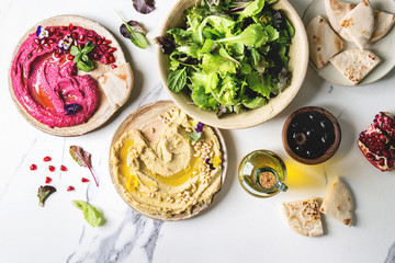 Variety of homemade traditional and beetroot spread hummus with pine nuts, olive oil, pomegranate served on ceramic plates with pita bread and green salad on white marble background. Flat lay, space. - obrazy, fototapety, plakaty