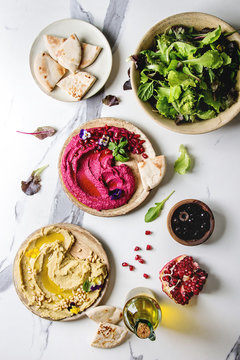 Fototapeta Variety of homemade traditional and beetroot spread hummus with pine nuts, olive oil, pomegranate served on ceramic plates with pita bread and green salad on white marble background. Flat lay, space.