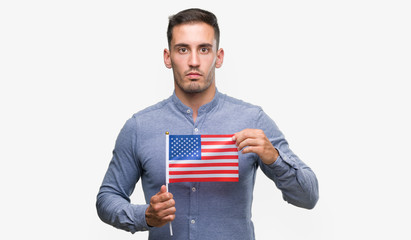 Handsome young man holding a flag of America with a confident expression on smart face thinking serious