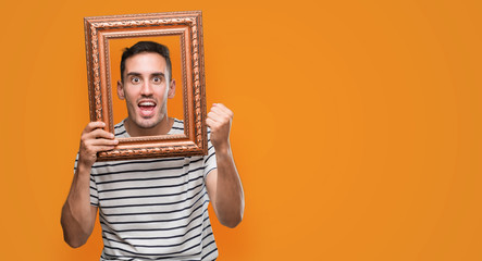 Handsome young man looking through vintage art frame screaming proud and celebrating victory and...