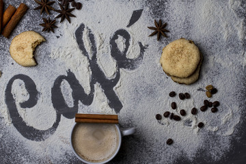 Dark table decorated with the word coffee made from wheat flour photographed from top to bottom, plus a large cup of fresh coffee and cinnamon stick with star anise and biscuits