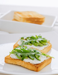 Roasted toasts with cream cheese and slices of avocado, leafs of rucola and pink Himalayan salt. Front view, low angle, focus on the centre of both breads. White wood background.