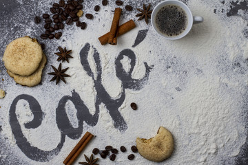 Dark table decorated with the word coffee made from wheat flour photographed from top to bottom, plus a small cup of fresh coffee and cinnamon stick with star anise and biscuits