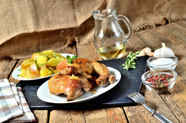 Golden grilled chicken legs on a plate and roasted potatoes, rosemary, jug with oil, oregano, salt, pepper and garlic on a wooden table