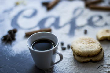 Dark table decorated with the word coffee made from wheat flour, plus a cup of fresh coffee in the foreground with selective focus and cinnamon stick with star anise and biscuits