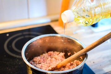 Sunflower oil is poured into a pot with fresh ground pork and a black and white onion on a glass ceramic plate. Afternoon preparing lunch in the kitchen. Wooden spoon in a pot.