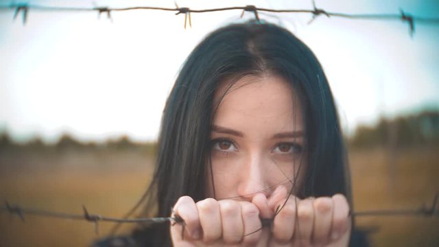 girl brunette refugee behind barbed wire camp slow motion video. lifestyle the concept of freedom is upset Woman hands and barbed wire. girl Refugee, prison, refugees captivity concept