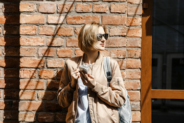 Girl with mobile phone in the city, near the house. Stylish woman with sunglasses with a briefcase. A young woman is waiting for a meeting. Selfy makes herself a beautiful lady.