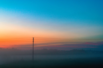 Evening thick fog over field and forest. Landscape of the evening sky with orange and blue sky and fog. Telegraph pole in the fog.