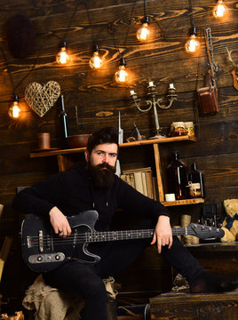 Guy in cozy warm atmosphere play relaxing soul music. Favourite activity. Man with beard holds black electric guitar. Man bearded musician enjoy evening with bass guitar, wooden background