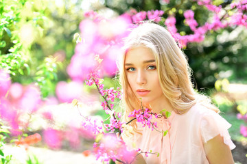 Fototapeta premium Portrait of lovely blond girl on natural background. Pretty young woman with blue eyes posing next to blooming flowers with pink petals, charm of spring concept
