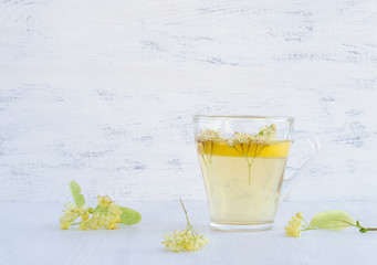A transparent Cup of tea with lemon and Linden flowers stands on a white wooden table. Linden flowers lie next to.