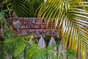 Humorous rustic wooden sign by picket fence in Key West surrounded by tropcal plants says Help a...