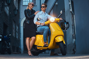 Fototapeta na wymiar Summer Europe vacation, date, romance. Attractive couple - handsome stylish guy dressed in a white shirt and jeans sitting on yellow classic italian scooter while his charming blonde girlfriend