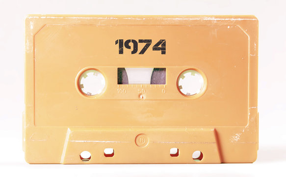 A vintage cassette tape from the 1980s era (obsolete music technology) with the text 1974 printed over it, stencil font. Color: cream, sand. White background.
