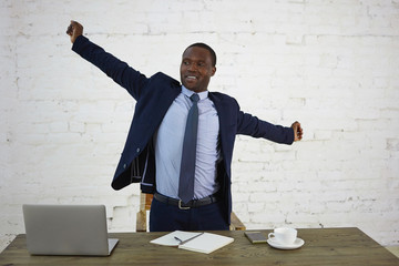 Handsome tired dark skinned businessman in elegant suit stretching arms, standing at desk with...