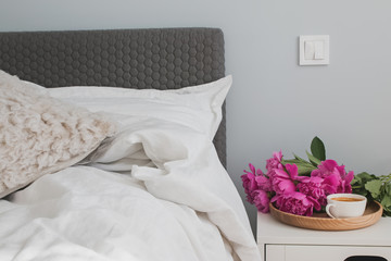 Bed with white bed sheets and pink peonies and coffee on the nightstand.