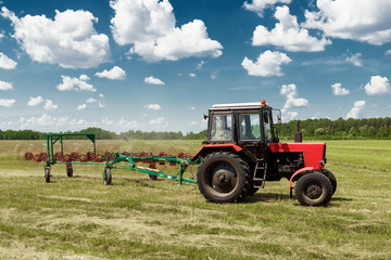 Agricultural machinery, a tractor collecting grass in a field against a blue sky. Hay harvesting, grass harvesting. Season harvesting, grass, agricultural land.