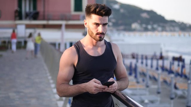 Athletic young man at the seaside using cell phone to type message while looking at the sea and beach