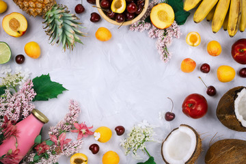 Flatlay frame arrangement with various organic fruits: bananas, coconuts, pineapple, peaches, fresh cherries, lilac flowers and smoothie bottle on grey cement background with copyspace