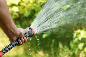 Men's hand with garden hose watering plants, gardening concept. Hand garden hose with water spray, watering flowers, close-up, water splashes, fresh green lawn. Copy space