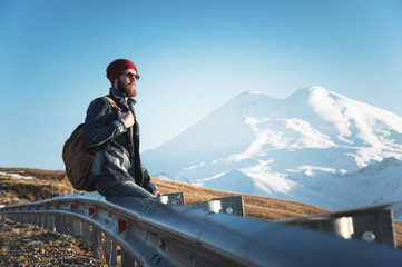 Bearded tourist hipster man in sunglasses with a backpack sitting on a roadside bump and watching the sunset against the background of a snow-capped mountain