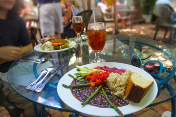 Vegetarian plate with black beans and rice and vegetables and cornbread on glass table with turquoise sunglasses and ice tea and another plate with a hamburger and blurred unrecognizable diners in bac