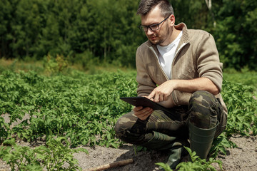 A male farmer sitting in the field and using a tablet. Modern application of technologies in agricultural activities. Farmland, new technologies, harvesting, fertilization, crop inspection.