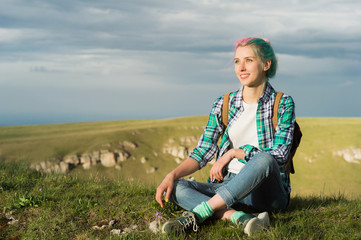 Naklejka na ściany i meble Portrait of young smiling woman traveler with multi-colored hair. Sitting high in the mountains in the evening with a decline in the background of a plateau