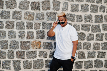 Stylish arabian muslim boy with originally hair and sunglasses posed on streets against brick wall.