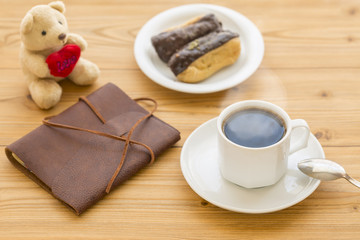 Hot coffee with steam and leather journal, teddy bear, and some eclairs