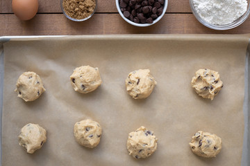 Flat lay of chocolate chip cookie dough balls on a baking sheet, with line of ingredient bowls