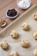 Close up on chocolate chip cookie dough balls on parchment paper, with ingredients in the background