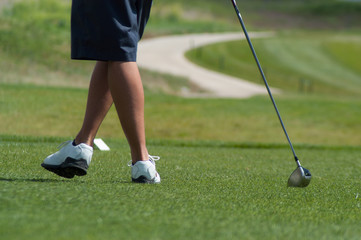 Young man leaning on his golf club on the putting green wearing blue shorts and white golf shoes. Playing golf in the summertime on a golf course.