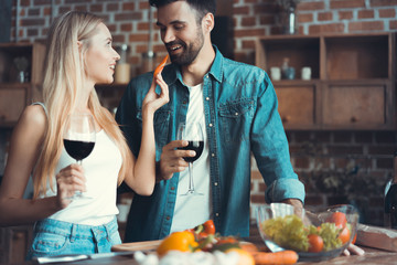 Beautiful young couple is feeding each other and smiling while cooking in kitchen at home.