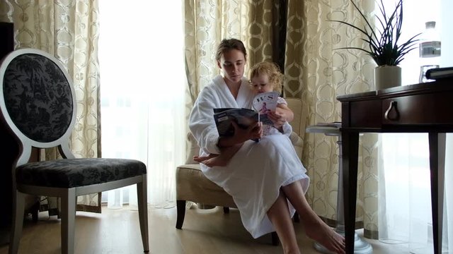 Mother and little girl sitting together in the armchair and reading magazine in the hotel room