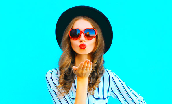 Cool girl with red lips is sends an air kiss in a sunglasses shape of heart over  blue background