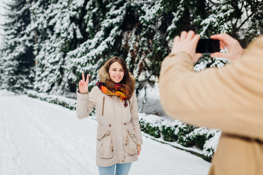 Young couple in love rest in the park winter. A man holds mobile phone in hands and takes a photo of girlfriend against the background of snowcovered trees. Woman in jacket and red backpack