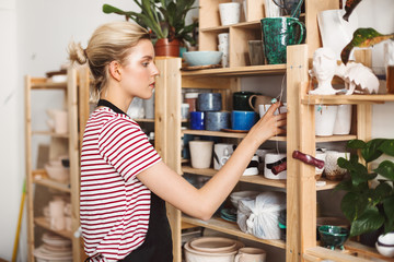 Beautiful girl in black apron and striped T-shirt thoughtfully putting handmade dishes on shelves at modern pottery studio