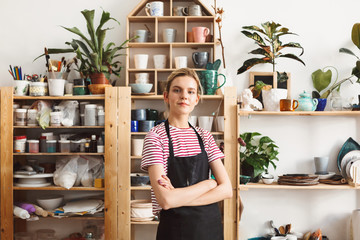 Beautiful girl in black apron and striped T-shirt dreamily looking in camera with handmade dishes on shelves on background at modern pottery studio