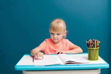 Portrait of little blonde girl is sitting at the table and paints. Schoolkid is doing her homework in classroom at school or at home. She shows finger on letters or numbers